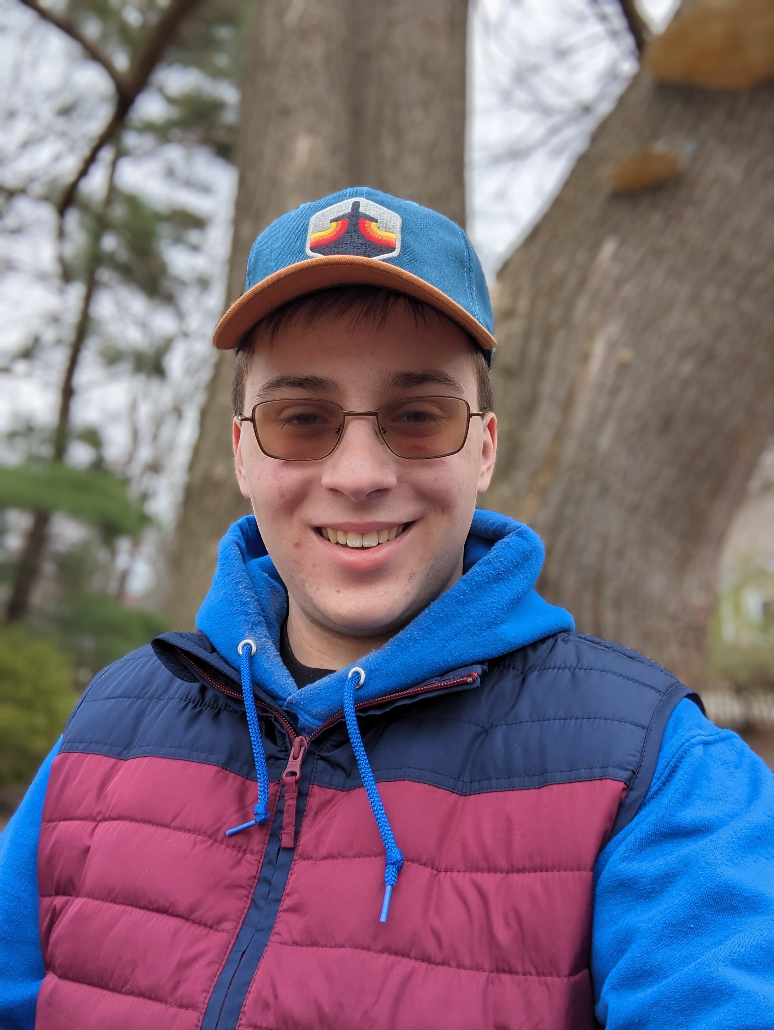 A photo of Matthew Brewer outside, up against a tree, wearing a blue hoodie and black t-shirt underneath a navy and red vest, and a blue and orange hat with a colored Jet Lag: The Game logo. The background is blurred, and Matthew's glasses are tinted brown.